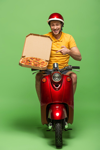 smiling delivery man in yellow uniform on scooter pointing with finger at pizza on green