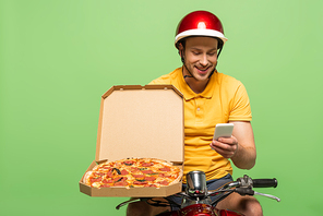 smiling delivery man in yellow uniform on scooter with pizza using smartphone isolated on green
