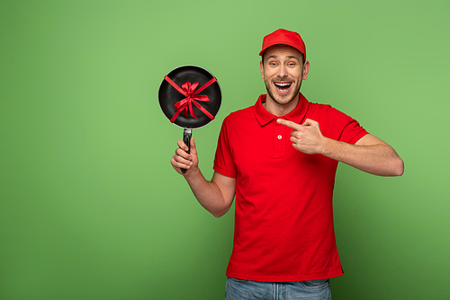 happy delivery man in red uniform pointing with finger at frying pan with bow on green