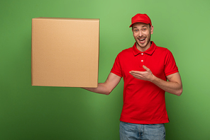 happy delivery man in red uniform pointing with hand at huge parcel on green