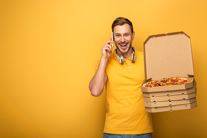 happy man with headphones in yellow outfit holding pizza and talking on smartphone on yellow background