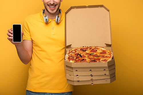 cropped view of happy man with headphones in yellow outfit holding pizza and smartphone on yellow background