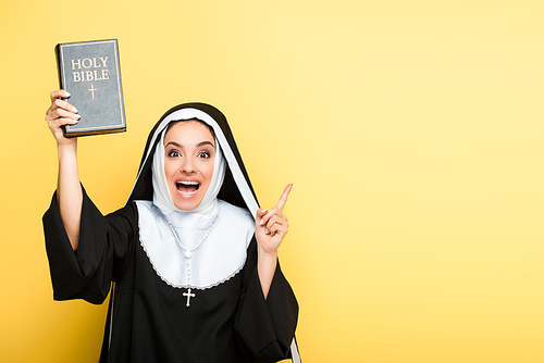 attractive excited nun holding holy bible and pointing up on grey