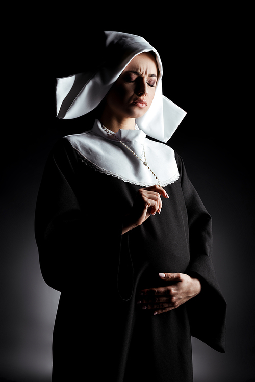 worried pregnant nun holding cross and touching belly on grey