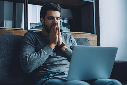 Shocked man with hands near mouth watching pornography on laptop in living room