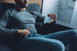 Cropped view of man masturbating while watching pornography on smartphone on sofa
