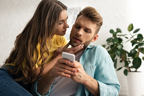 Attractive girl touching boyfriend with smartphone in living room