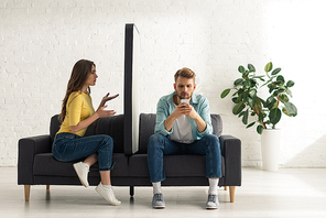 Confused girl looking at big model of smartphone near addicted boyfriend chatting on sofa