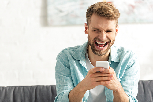 Selective focus of cheerful man laughing while using smartphone on couch
