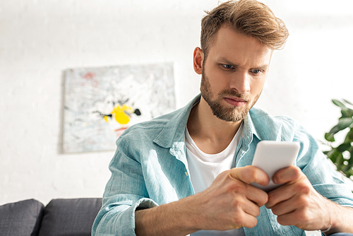 Selective focus of angry man using smartphone on sofa in living room
