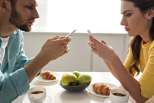 Side view of couple using smartphones near coffee and croissants on table