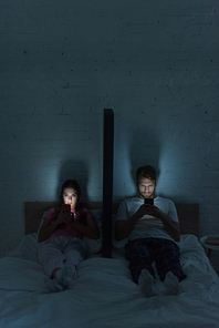 Young couple with smartphones sitting near model of smartphone on bed at night