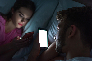 Selective focus of man using smartphone near girlfriend chatting on bed at night