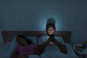 Smartphone depended man sitting on bed near sleeping girlfriend