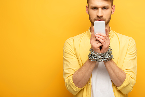Handsome man with metal chain around hands holding smartphone on yellow background