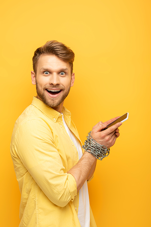 Side view of cheerful man with metal chain around hands holding smartphone and  on yellow background