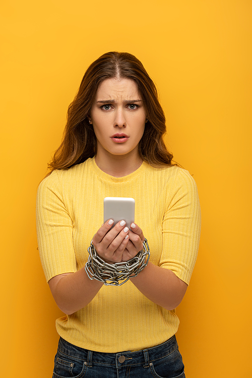 Confused girl  while holding smartphone in tied hands with metal chain on yellow background