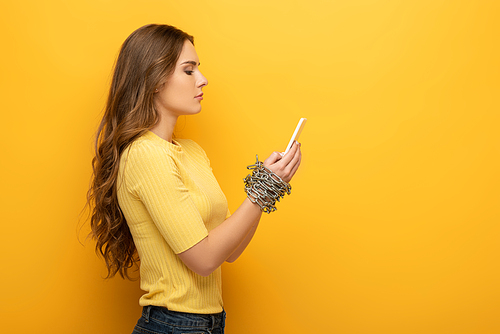 Side view of attractive woman holding smartphone in tied hands with metal chain on yellow background