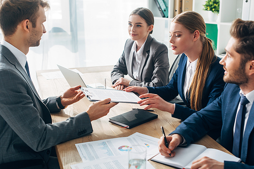 Side view of employee holding clipboard with resume near recruiters at table