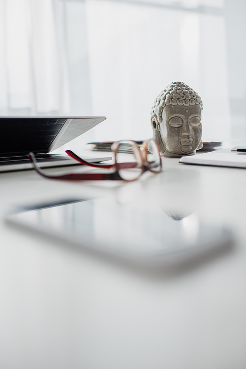 selective focus of Buddha head, eyeglasses and laptop on table in modern office