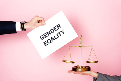 cropped view of businessman holding placard with gender equality lettering near businesswoman with golden scales on pink, gender equality concept