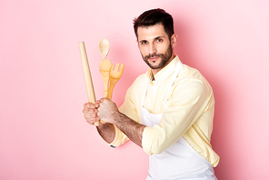 handsome man in apron  while holding wooden rolling pin, spoon and fork on pink