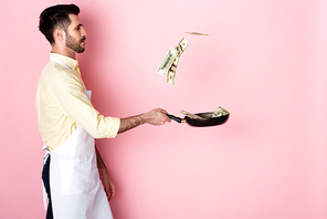 side view of bearded man in apron holding frying pan with money on pink