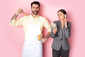 handsome man in apron holding wooden rolling pin near businesswoman with money talking on smartphone on pink