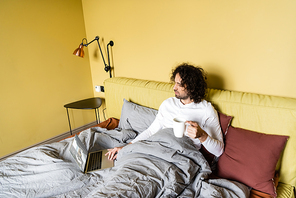 high angle view of young freelancer using laptop with tickets online website while holding cup of coffee in bed