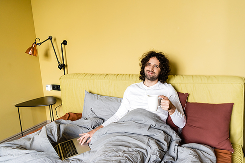 happy freelancer smiling at camera while using laptop and holding cup of coffee in bed
