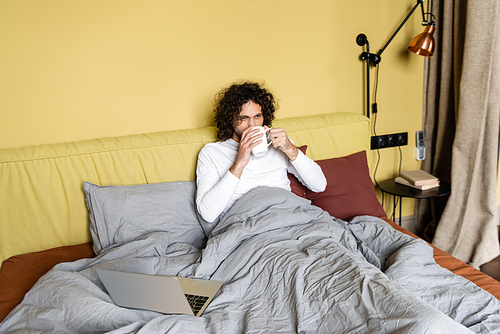 curly freelancer drinking coffee in bed near laptop and looking away