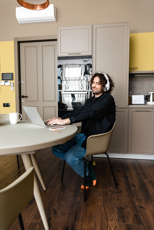 happy freelancer in wireless headphones using laptop in kitchen