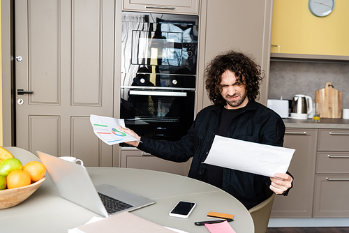 Selective focus of confused freelancer holding papers with charts near gadgets on kitchen table