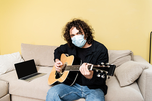 Man in medical mask playing acoustic guitar near laptop on sofa