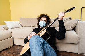 Curly man in medical mask playing acoustic guitar while sitting on floor at home