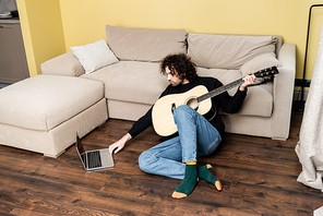 Handsome man holding acoustic guitar and using laptop on floor at home