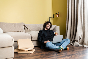 Selective focus of smiling man looking at laptop and holding bottle of beer near pizza boxes on floor at home