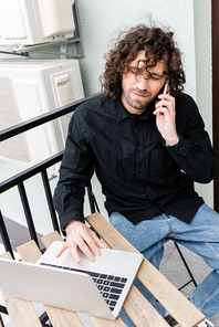 Selective focus of handsome man talking on smartphone and using laptop on balcony at home