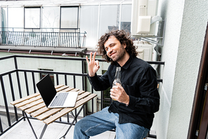 Freelancer smiling at camera and showing ok gesture while holding beer bottle near laptop on balcony