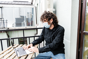 Freelancer in medical mask using laptop on balcony at home