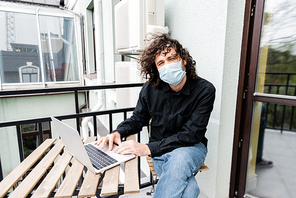 Man in medical mask using laptop on table on balcony