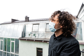 Side view of curly man in medical mask looking away while standing on balcony