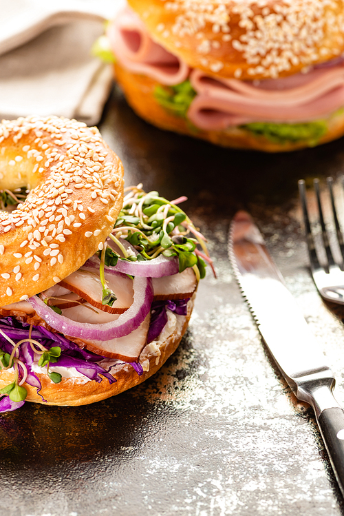 selective focus of fresh delicious bagel with meat, red onion, cream cheese and sprouts near cutlery on textured surface