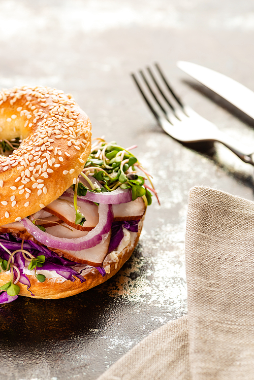 selective focus of fresh delicious bagel with meat, red onion, cream cheese and sprouts near napkin with cutlery on textured surface
