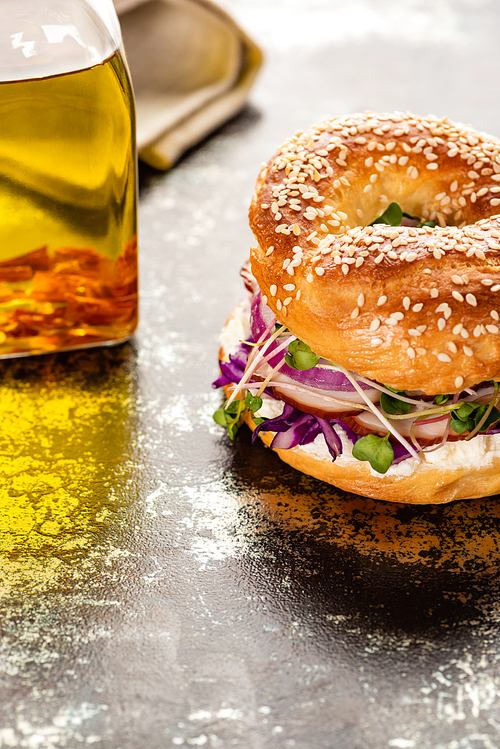 fresh delicious bagel with meat, red onion, cream cheese and sprouts on textured surface with bottle of oil