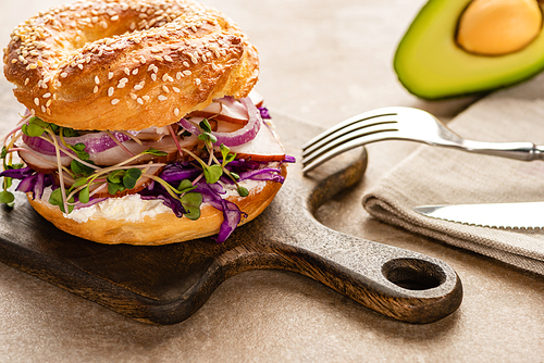 fresh delicious bagel with meat, red onion, cream cheese and sprouts on wooden cutting board near napkin and cutlery