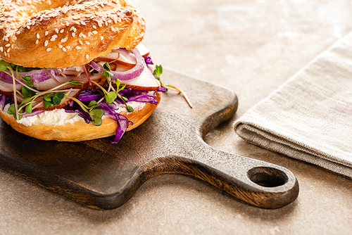 fresh delicious bagel with meat, red onion, cream cheese and sprouts on wooden cutting board near napkin
