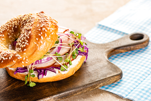 fresh delicious bagel with meat, red onion, cream cheese and sprouts on wooden cutting board near plaid napkin