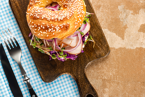 top view of fresh delicious bagel with meat, red onion and sprouts on wooden cutting board and plaid blue napkin with cutlery