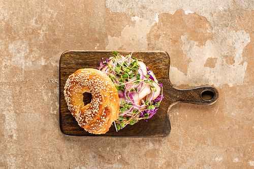 top view of fresh delicious bagel with meat, red onion and sprouts on wooden cutting board on aged beige surface